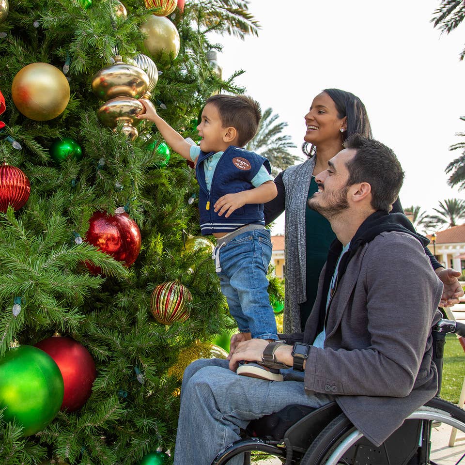 Author, Danny Pitaluga (right) sits in his black wheelchair wearing a charcoal hoodie as his wife, Val (middle), and his son, Joey (left), stands on Danny's lap to decorate a Christmas tree outdoors.