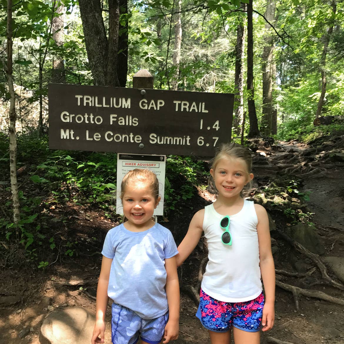 Author, Chris Johnstons' daughters, Kyndall (right), and Kyler (left) pose with a brown, wooden sign that reads, 'Trillium Gap Trail, Grotto Falls 1.4, Mt. LeConte, Summit 6.7.'