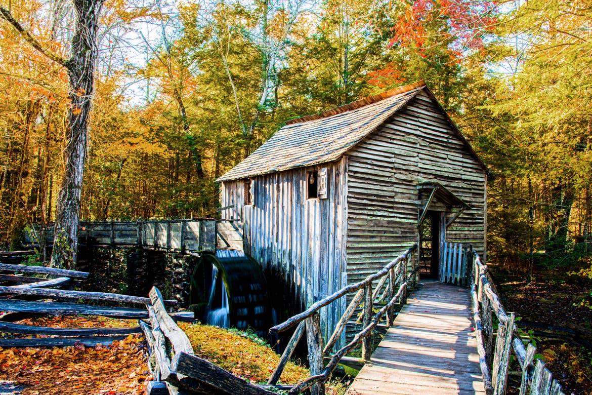 A wooden mill in the middle of a forest surrounded by fall foliage.
