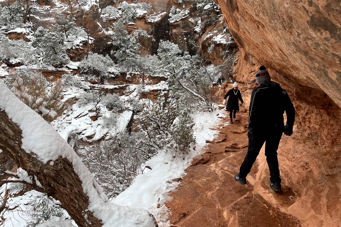 A man wearing black pants, jacket and a grey knitted cap walks along a trail where a woman in black pants, jacket and cap are surrounded by snowcapped red rock formations.