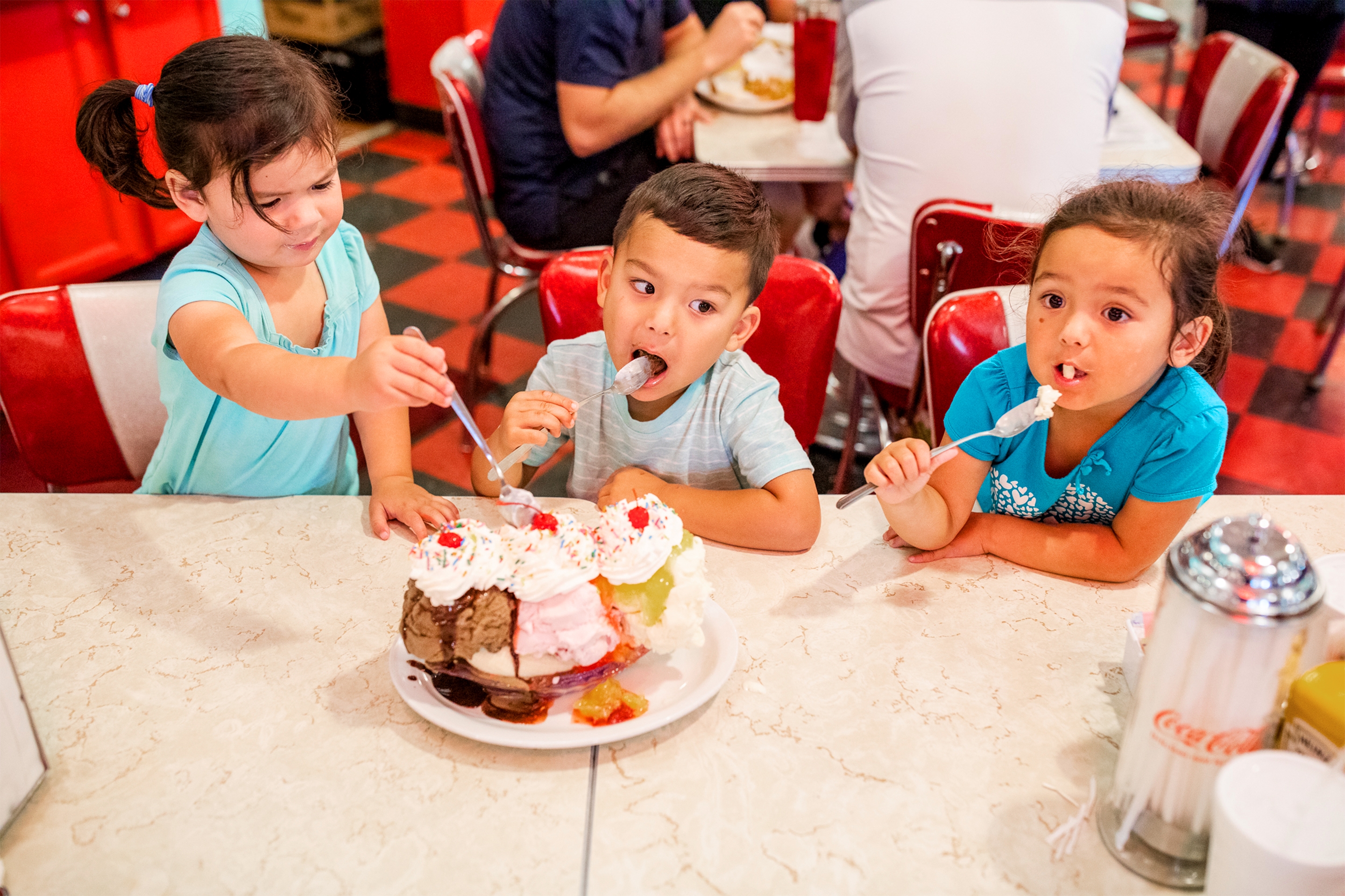Three Asian Pacific Islander children sit at a table enjoying a large ice cream sundae in a diner.