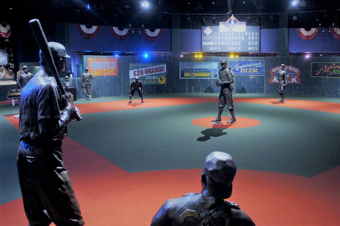 Several bronze statues resembling baseball players stand on a scaled indoor baseball field.