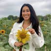 A woman stands in a sunflower field wearing a white denim jacket and holding a sunflower.