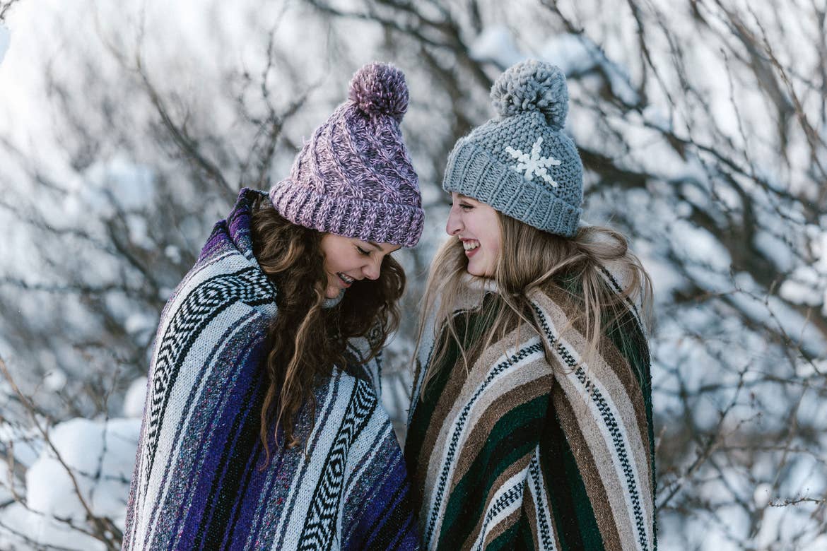 Two girls outside in the snow wearing blankets and beanies while laughing.