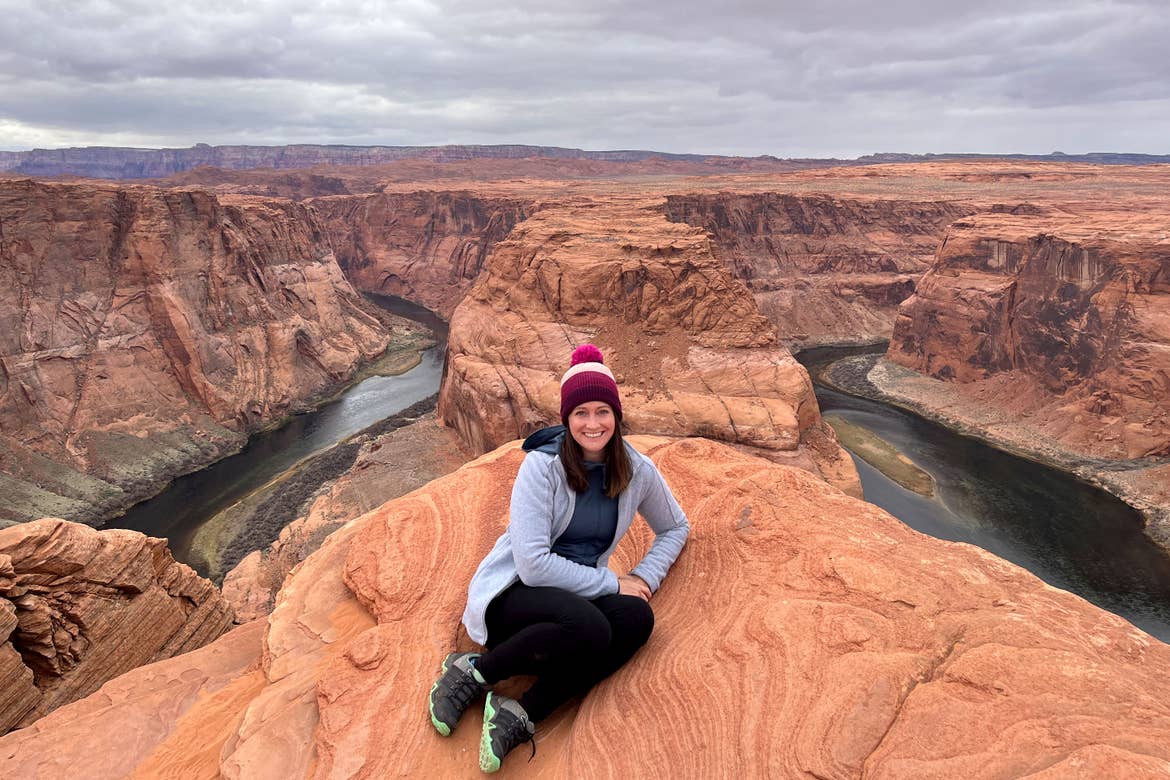 A woman in a magenta knitted hat, grey fleece jacket, aqua pullover and black tights sits on a red rock formation overlooking the Horseshoe Bend.