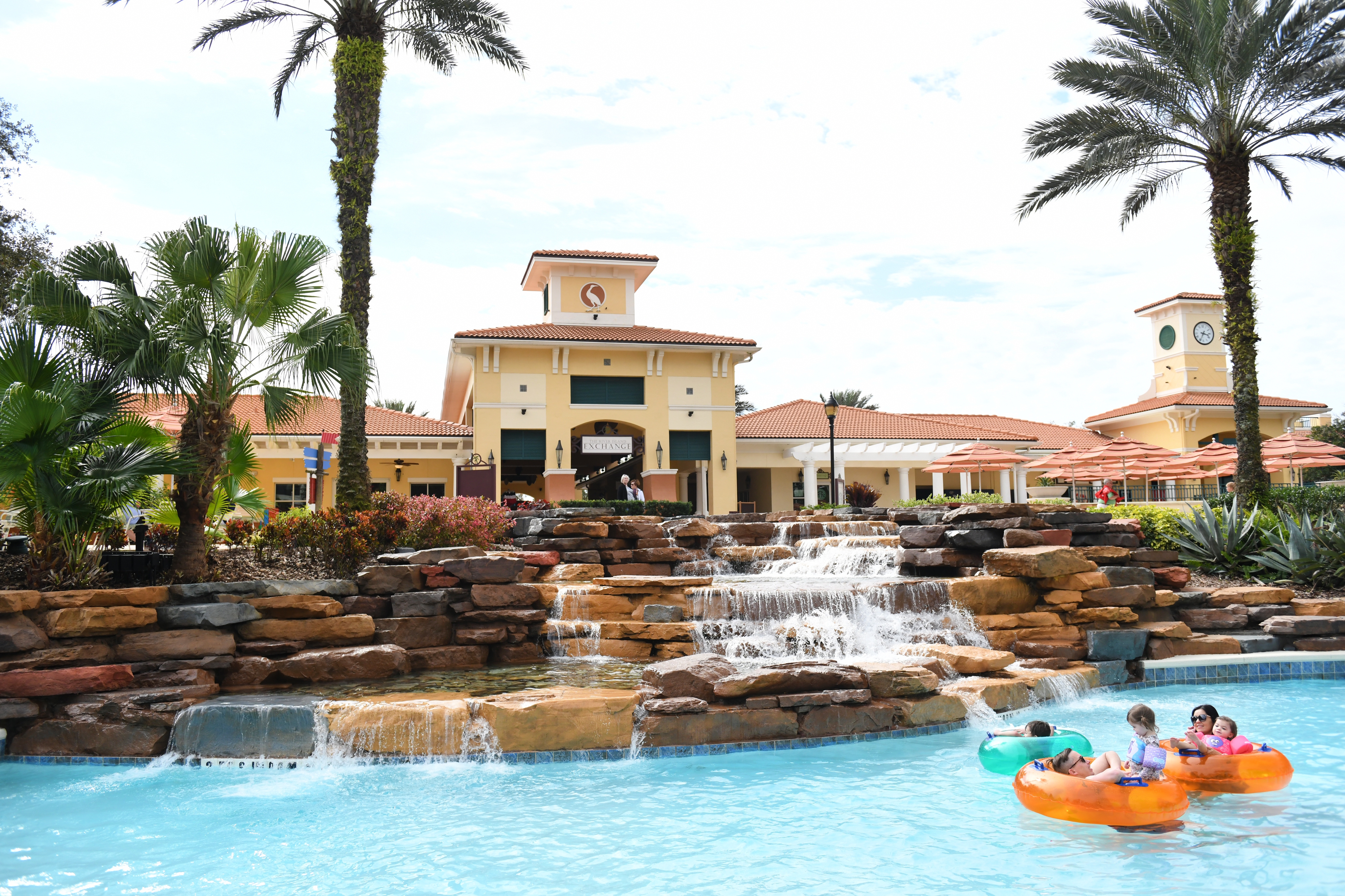a woman, man and two children float on orange and blue inner-tubes down a lazy river at an outdoor resort.