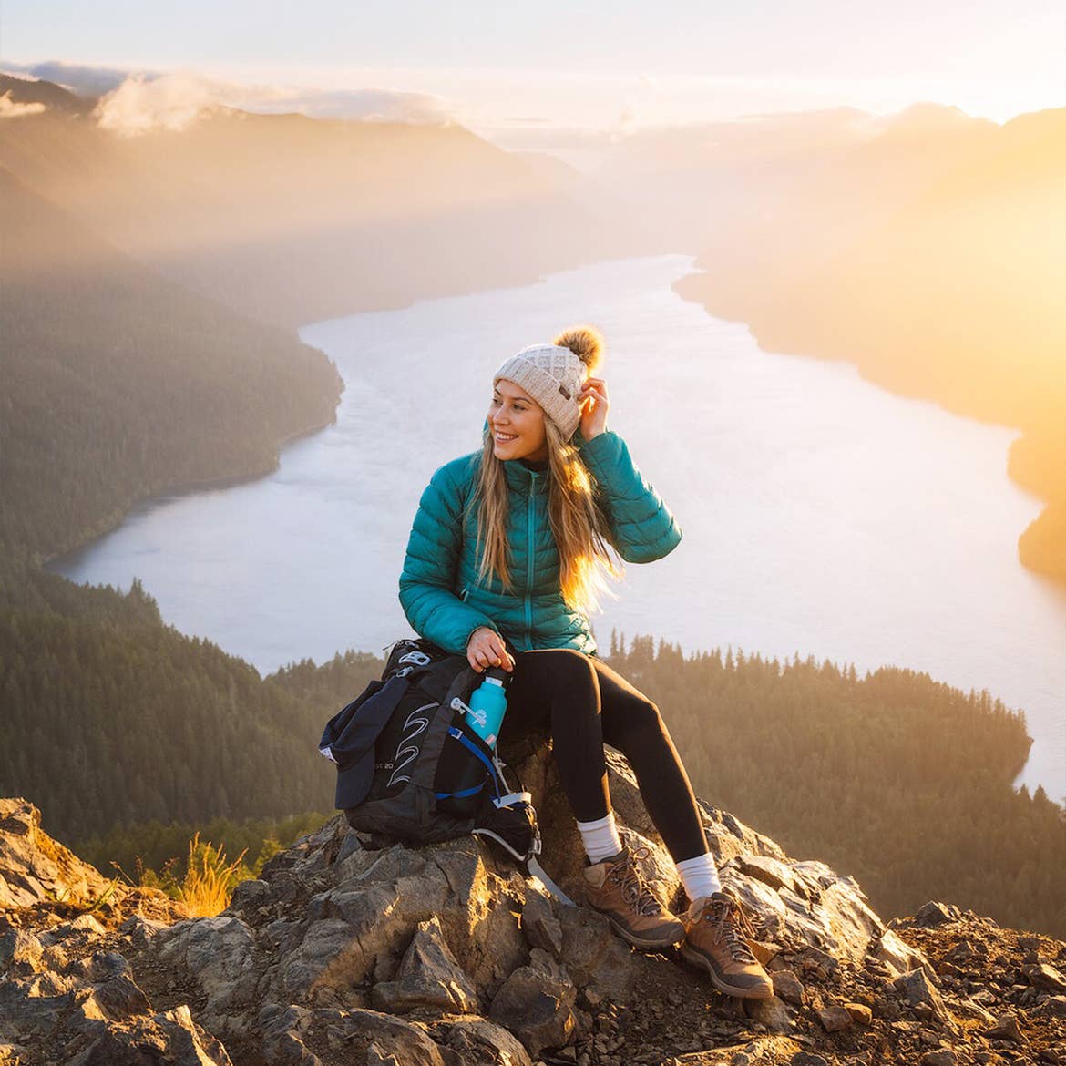 A caucasian woman wearing a teal winter coat, white beanie, black leggings and brown boots sits on a rock at a high elevation above a lake surrounded by trees and mountains.