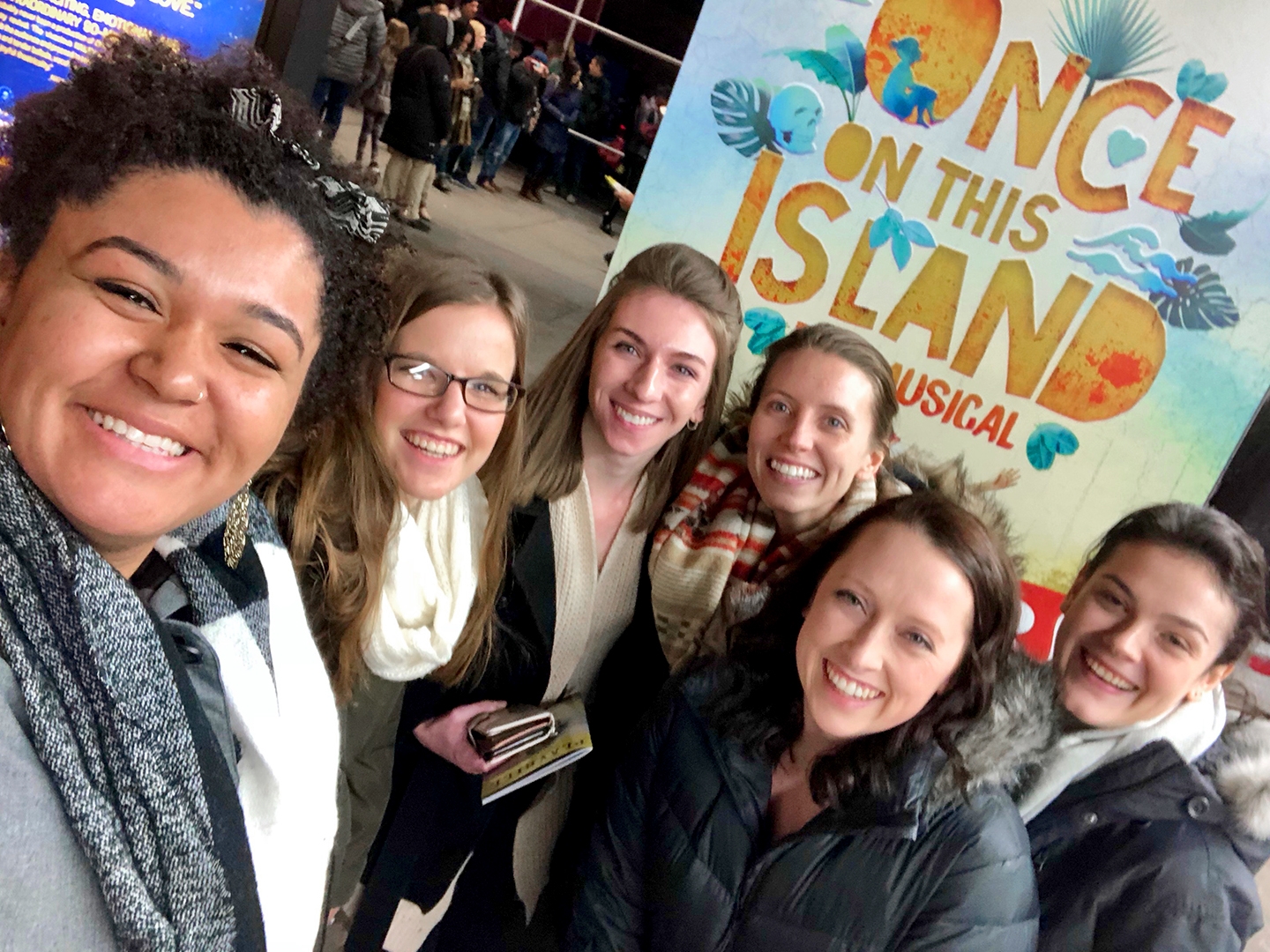 Several women stand wearing scarves and winter coats in front of a sign that reads, 'Once On This Island - A Musical' in New York City.