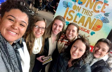 Several women stand wearing scarves and winter coats in front of a sign that reads, 'Once On This Island - A Musical' in New York City.