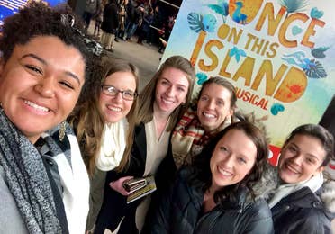 Several women stand wearing scarves and winter coats in front of a sign that reads, 'Once On This Island - A Musical' in New York City.