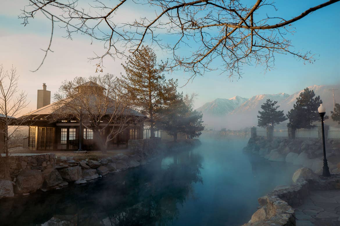 A brick hut stands near a hot spring and mountain range as steam and fog arise.