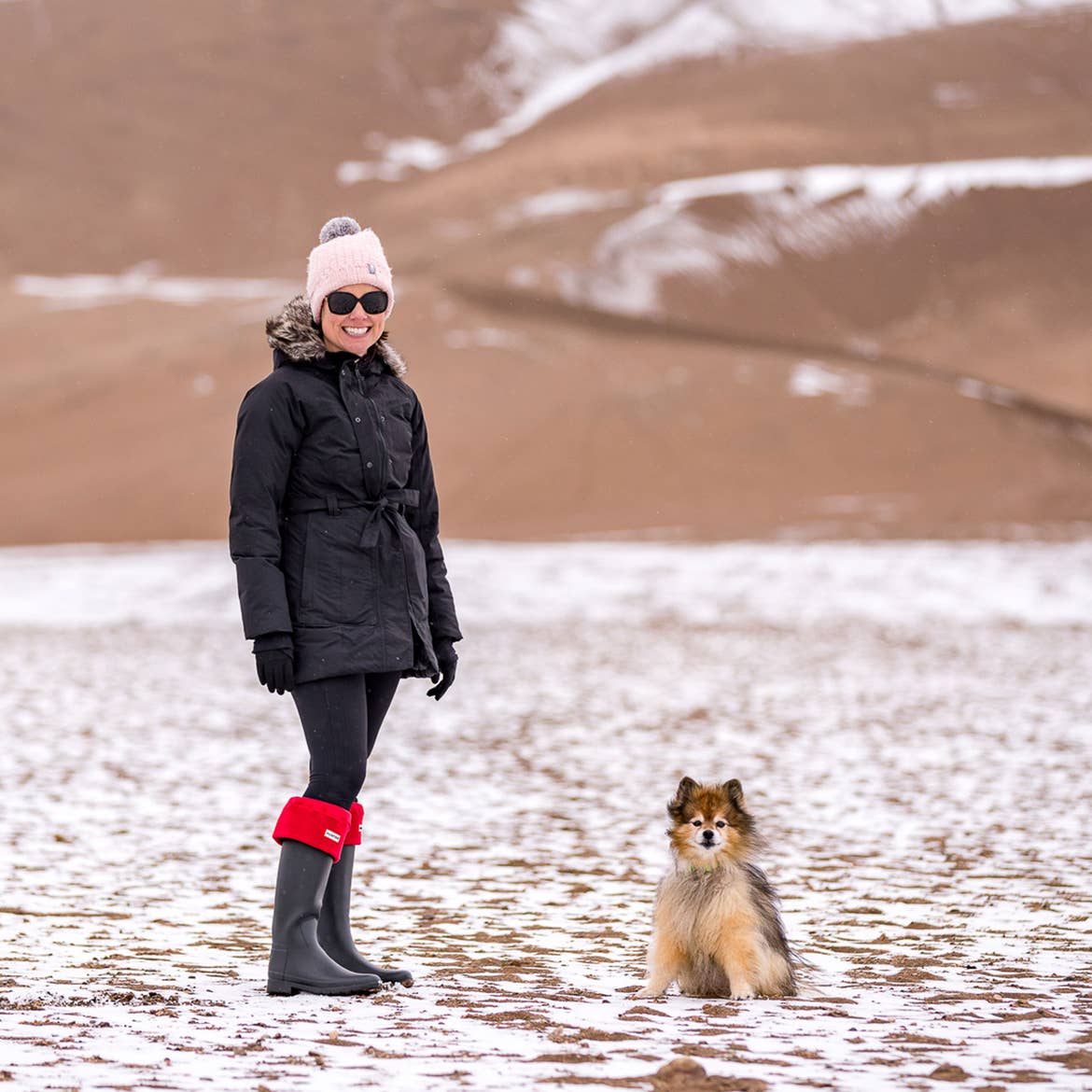 Author, Lauren Layne, stands in winter apparel with Bailey the Pomeranian at the Great Sand Dunes National Park and Preserve.