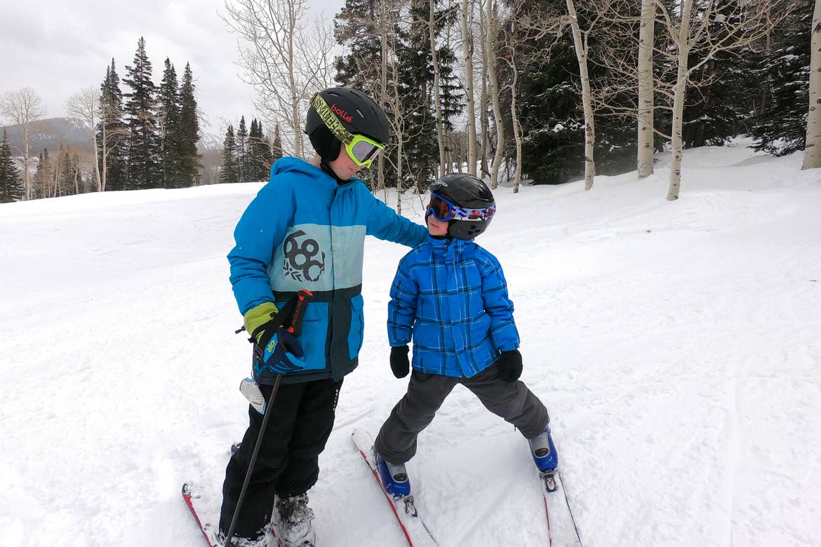 A young boy puts his arm around a smaller boy as they both wear black safety helmets, snow goggles, and blue winter apparel with skis on a snowy slope.