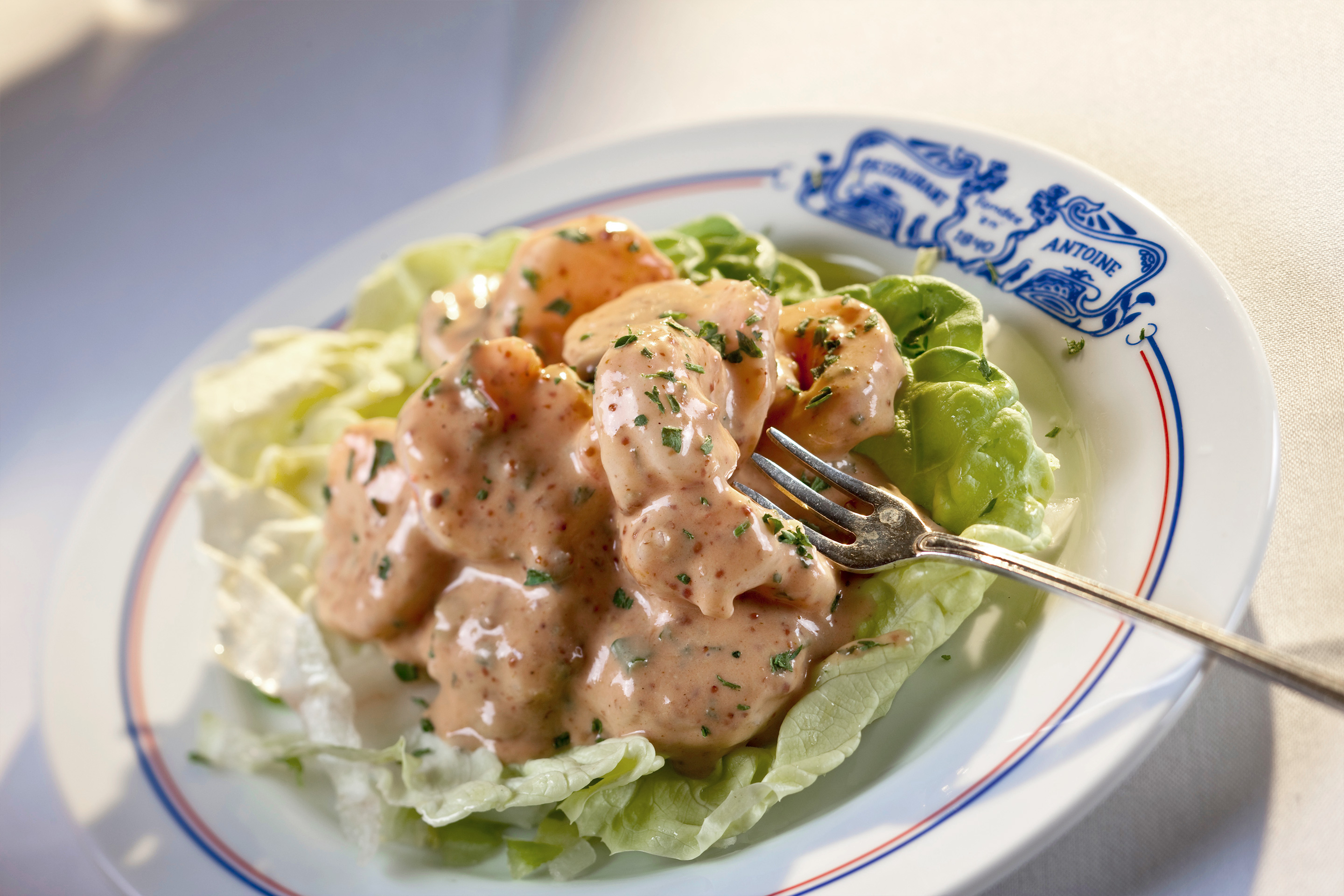 A white plate with blue toile imprint that reads 'Antoines' containing shrimp in Antoine's rémoulade.