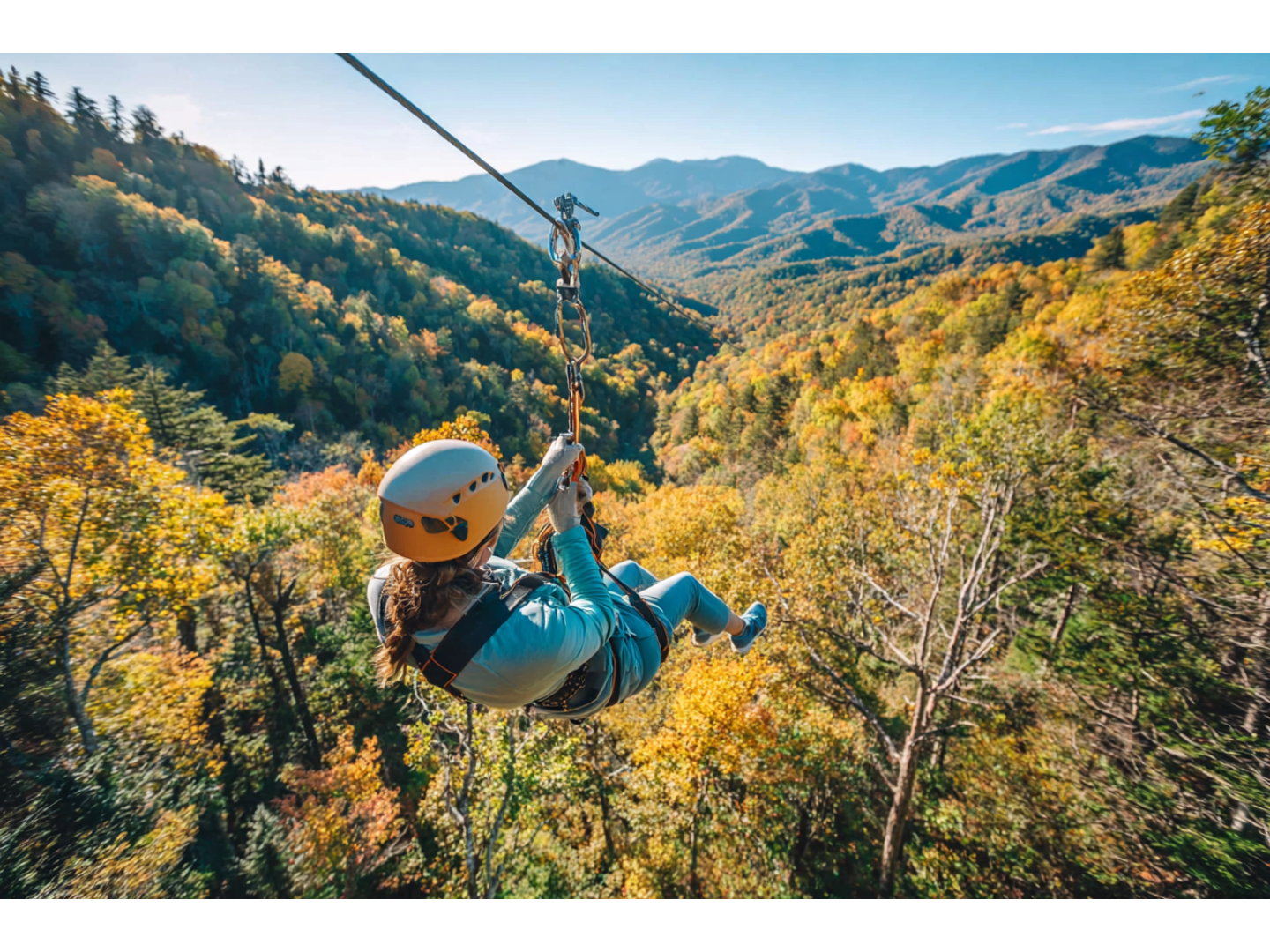 Person ziplining over colorful autumn forest with distant mountain range views.