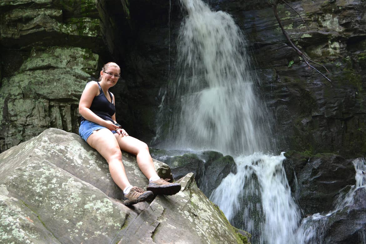 A woman wearing a purple tank top, jean shorts and glasses poses near waterfall cascades over a rock formation at Baskins Creek Trailhead in Tennessee.
