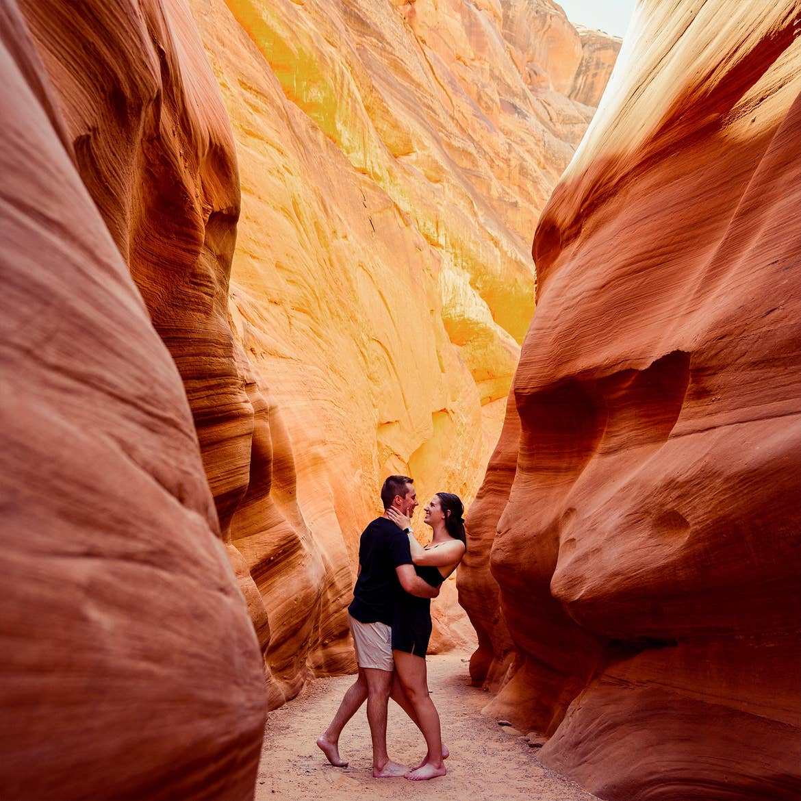 A man and woman wearing black outfits embrace in a red rock formation.