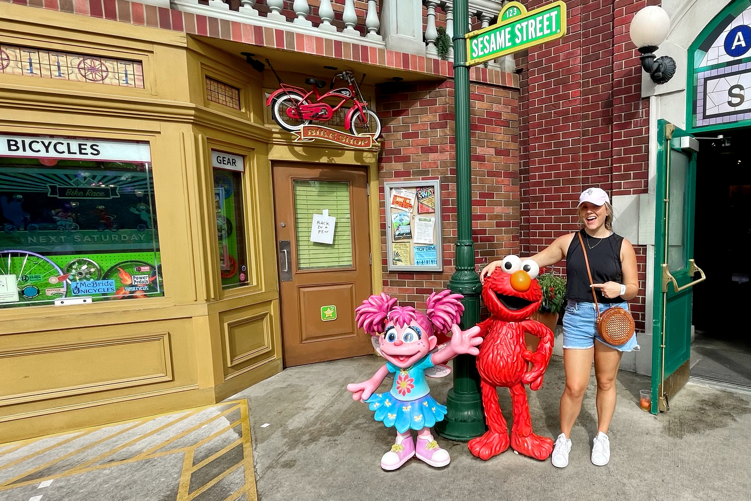 A Caucasian woman with a pink baseball cap, black tank, denim shorts and white shoes stands next to statues of Elmo and Abby Cadabby at Sesame Street Land™ in SeaWorld Orlando.