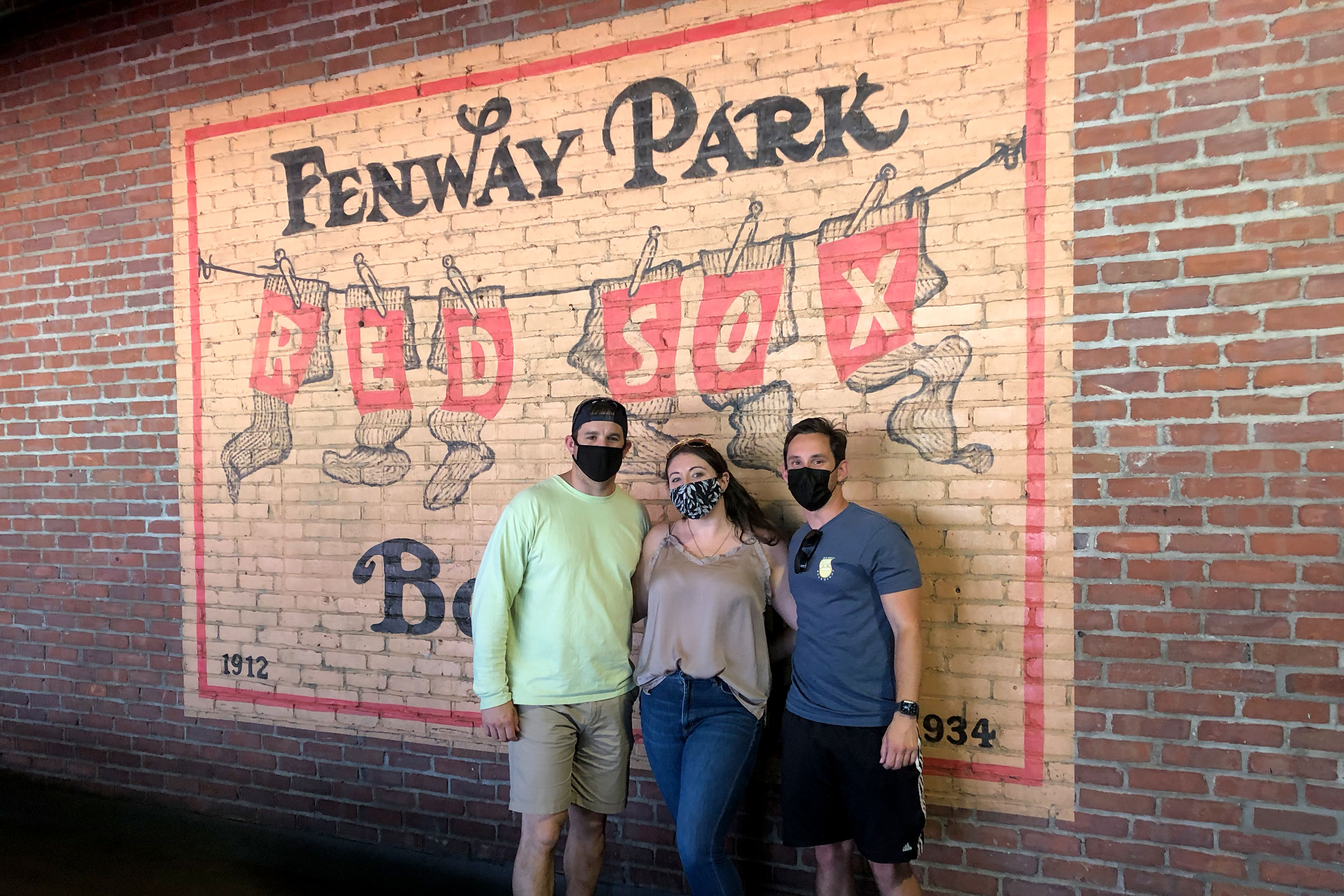 From left to right: A man, woman and man stand wearing safety masks in front of a Fenway Park wall mural.