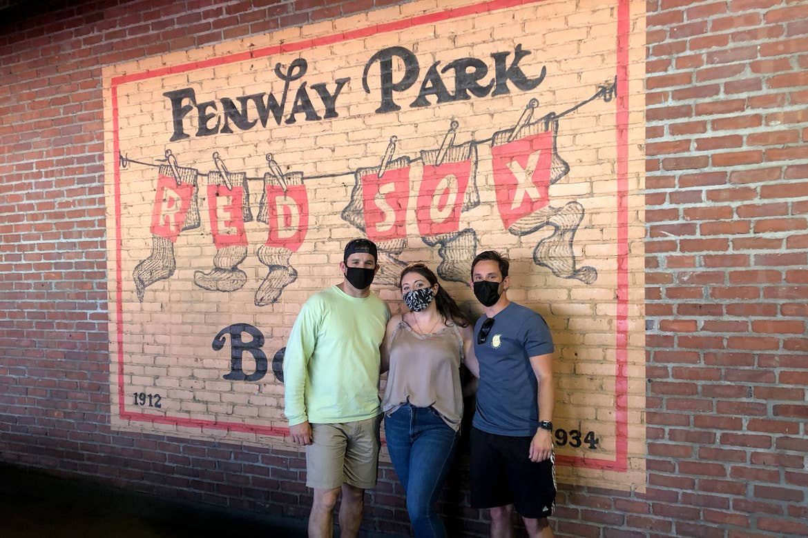 From left to right: A man, woman and man stand wearing safety masks in front of a Fenway Park wall mural.