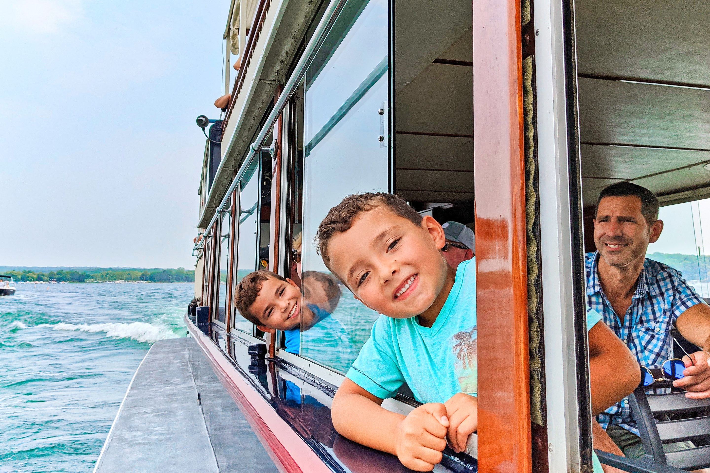 Two young boys and a man sit in a mailboat crossing a river.