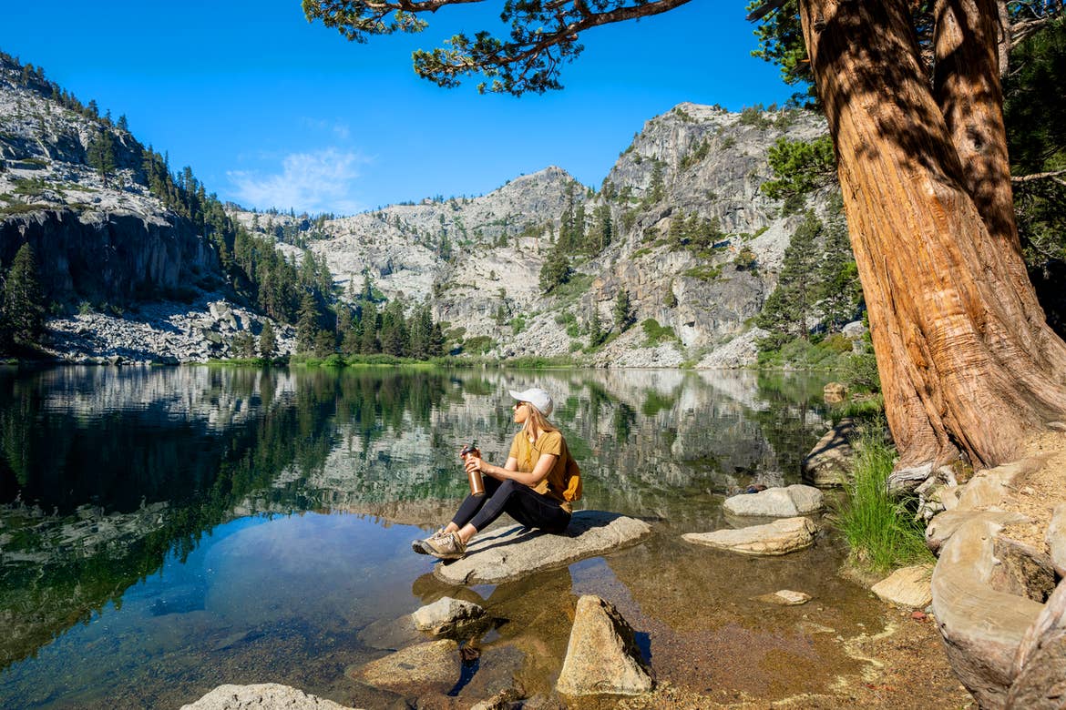 A caucasian woman wearing a mustard shirt, white baseball cap, black leggings and boots sits on a rock near a lake surrounded by pine trees on a rocky mountainscape.