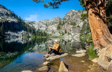 A caucasian woman wearing a mustard shirt, white baseball cap, black leggings and boots sits on a rock near a lake surrounded by pine trees on a rocky mountainscape.