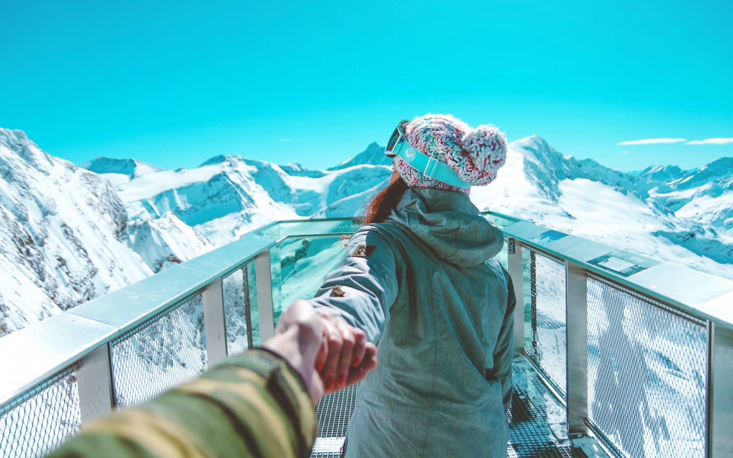 A woman holds a man's hand while wearing winter apparel and ski goggles looking off at snowcapped mountains under a blue sky.
