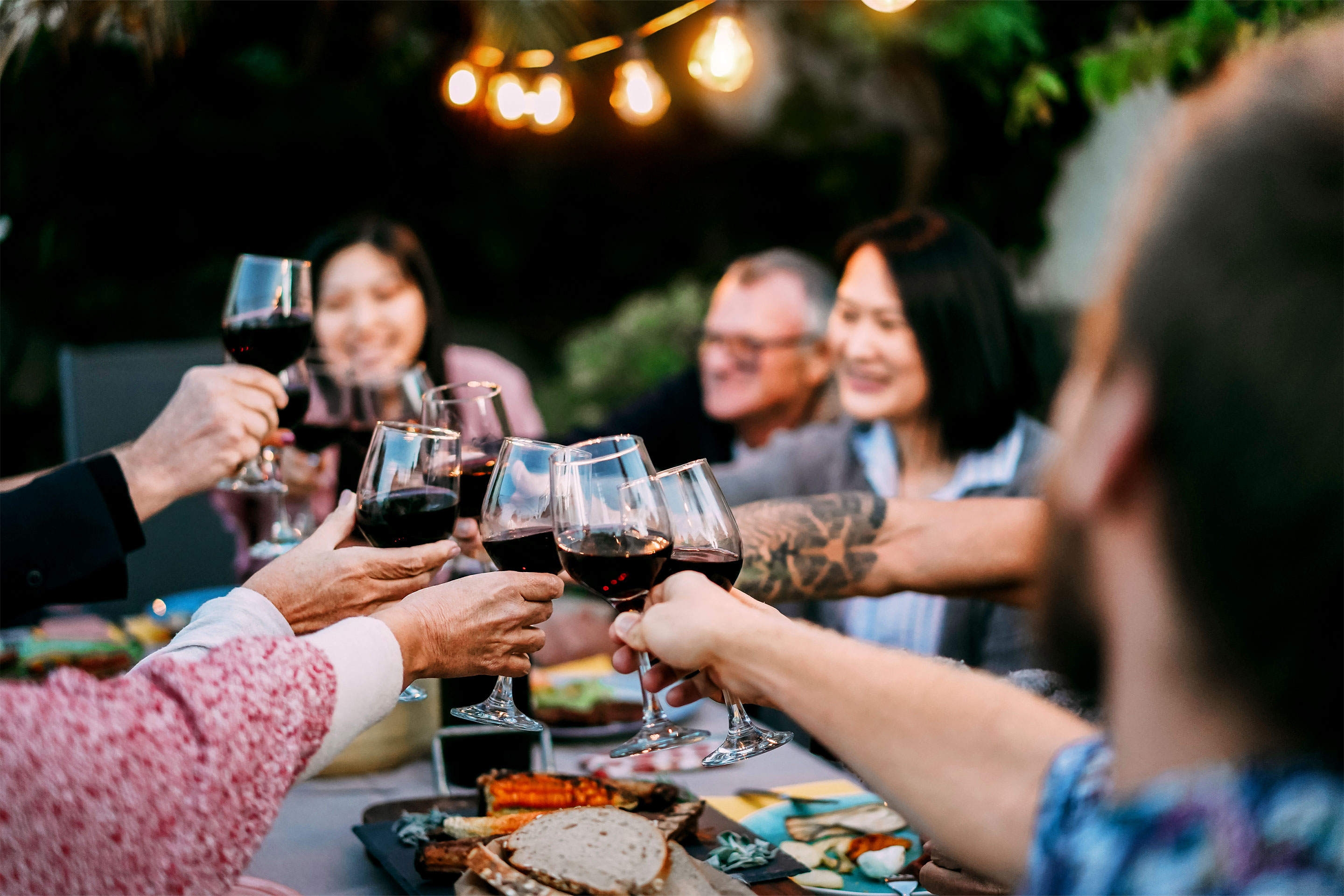 Several guests sit under string lights and an outdoor patio table giving 'cheers' with wine glasses.