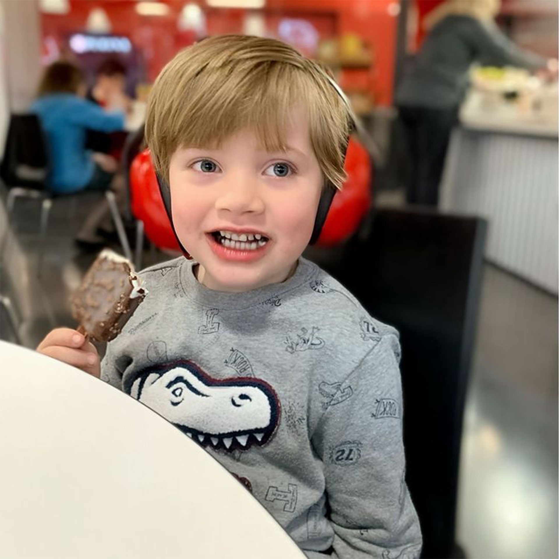Featured Contributor, Alicia Trautwein's son eats an ice cream bar while wearing red headphones at a dining table indoors.