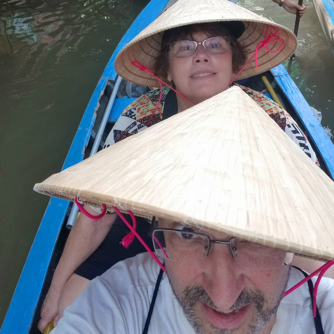 A caucasian man (front) and a caucasian woman (back) wear Vietnamese straw hats on a blue passenger kayak on the Mekong Delta in Vietnam.