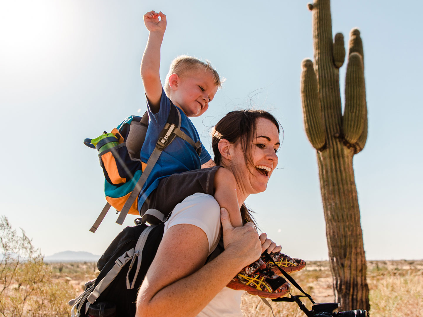 Jessica and her son laughing together while he sits on her shoulders as they hike in the Arizona desert.