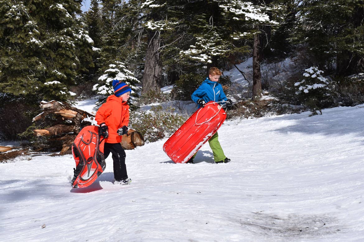 A two boys walk up a snowy hill holding sleds while wearing winter apparel.