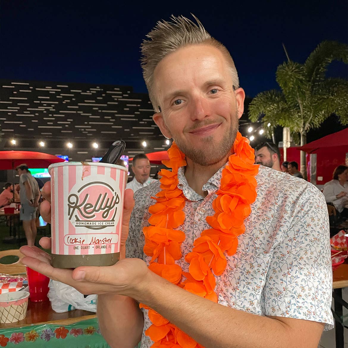 A man in a button-up shirt and orange lei holds a pint of pink and white striped ice cream outdoors.