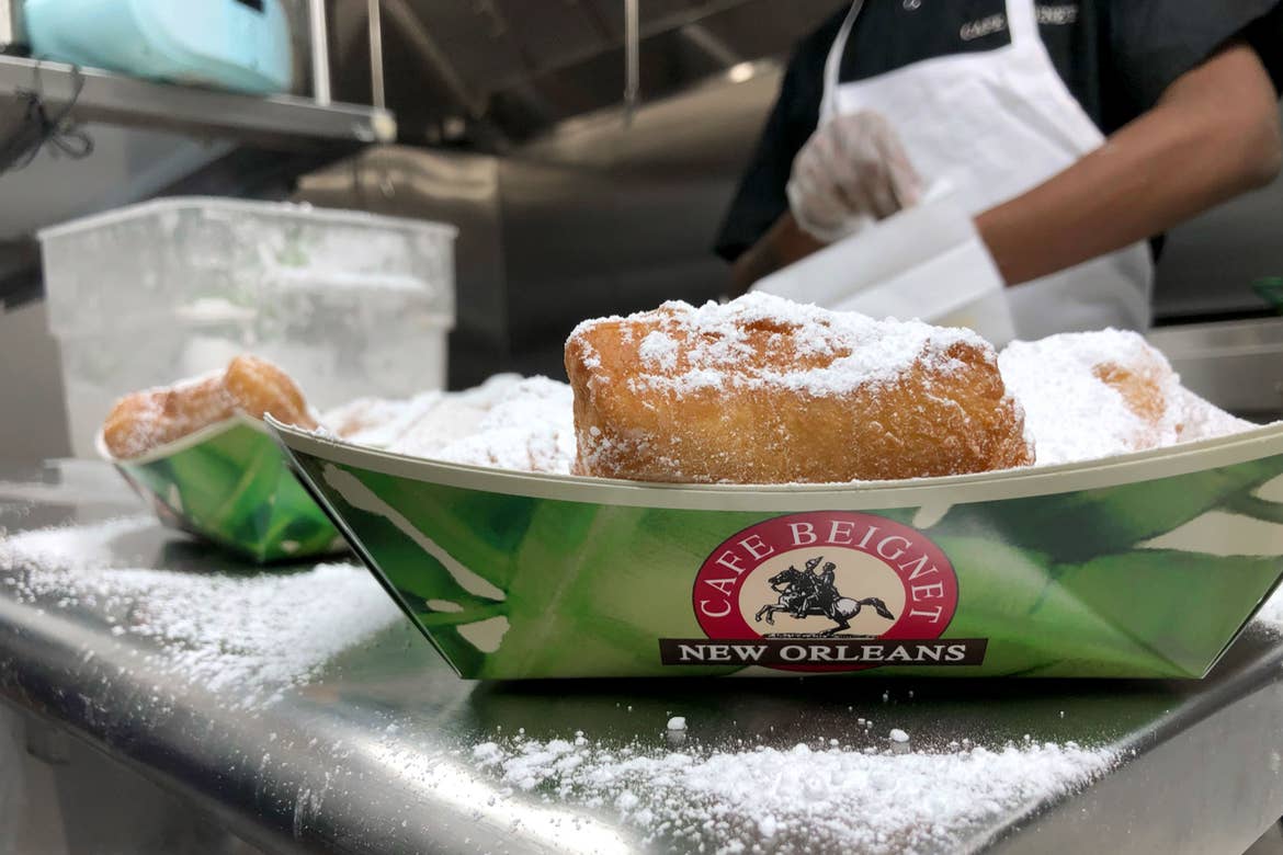 A pile of beignets in a green takeaway container that reads, 'Cafe Beignet, New Orleans' covered in powdered sugar in front of a baker.