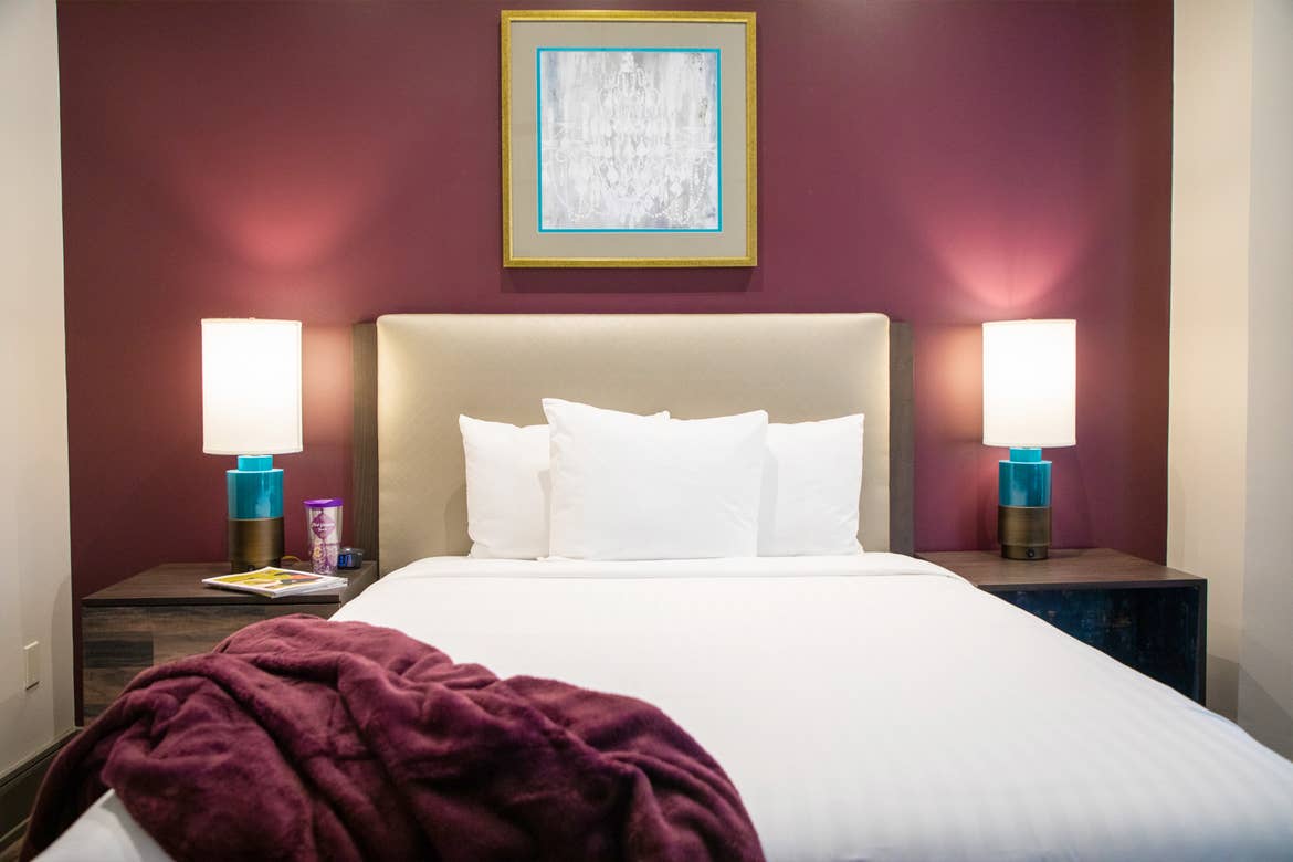 The master bedroom of our one-bedroom standard villa at our New Orleans resort furnished with a queen-sized bed with white sheets, two lamps and nightstands, a purple feature wall.