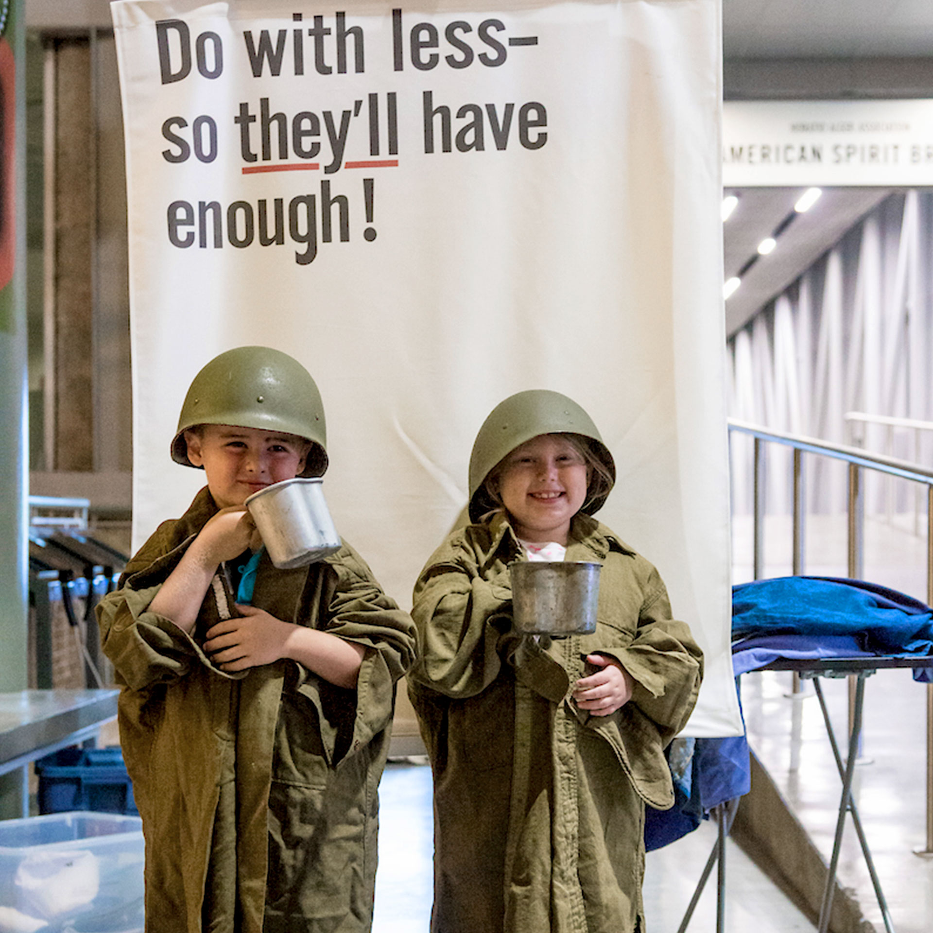 Two children recreate WWII propaganda dressed in 'Army Greens' and a helmet while holding silver mugs in front of a sign that reads, 'Do with less- So they have enough!'
