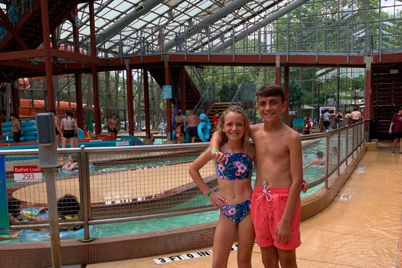 A caucasian girl (left) and boy (right) stand in swimsuits near an indoor waterpark.