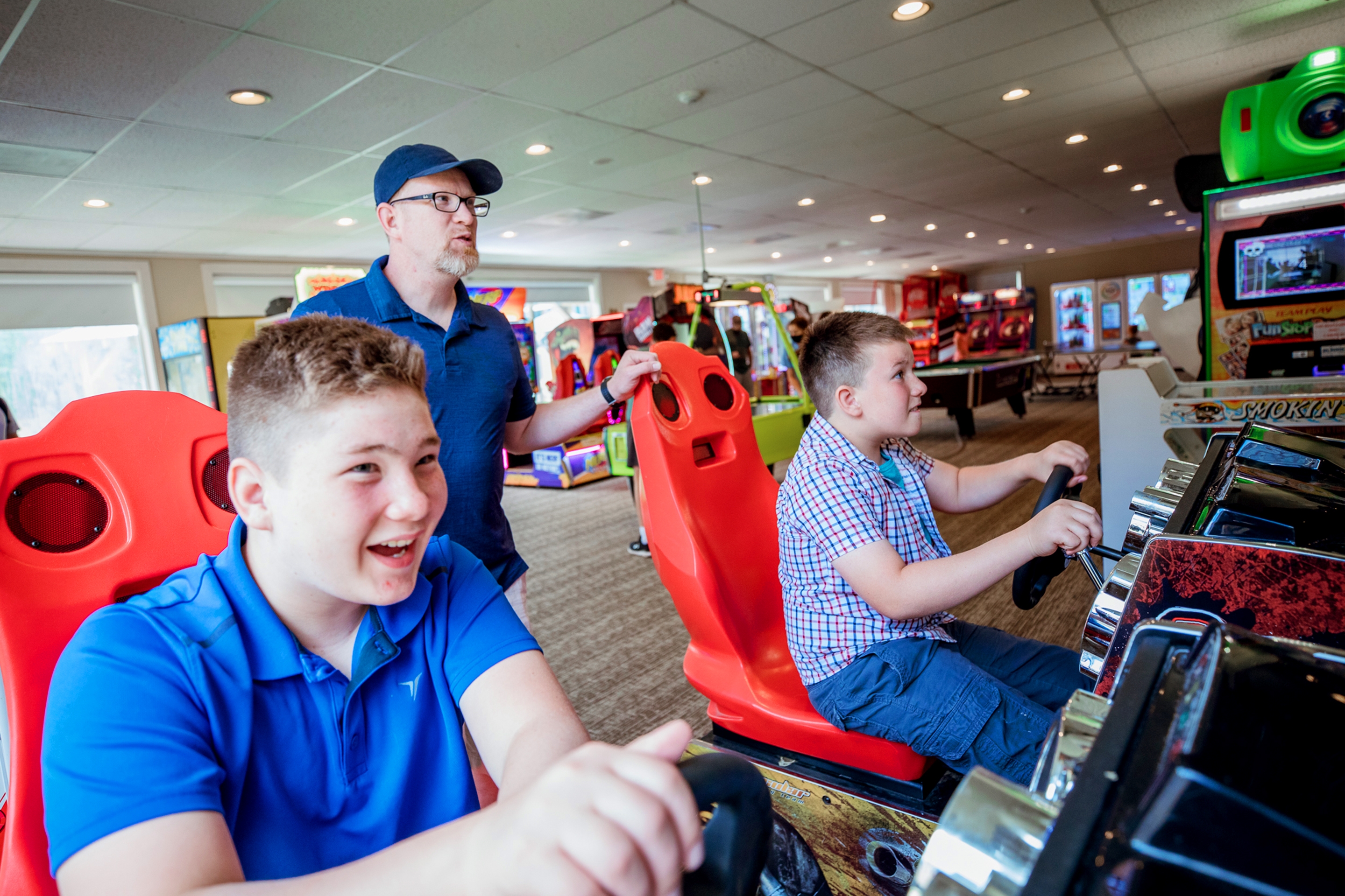 A caucasian man (middle) wearing a navy baseball cap and polo stands behind two tween, caucasian boys (left and right) seated in an arcade racing game seat behind a steering wheel.