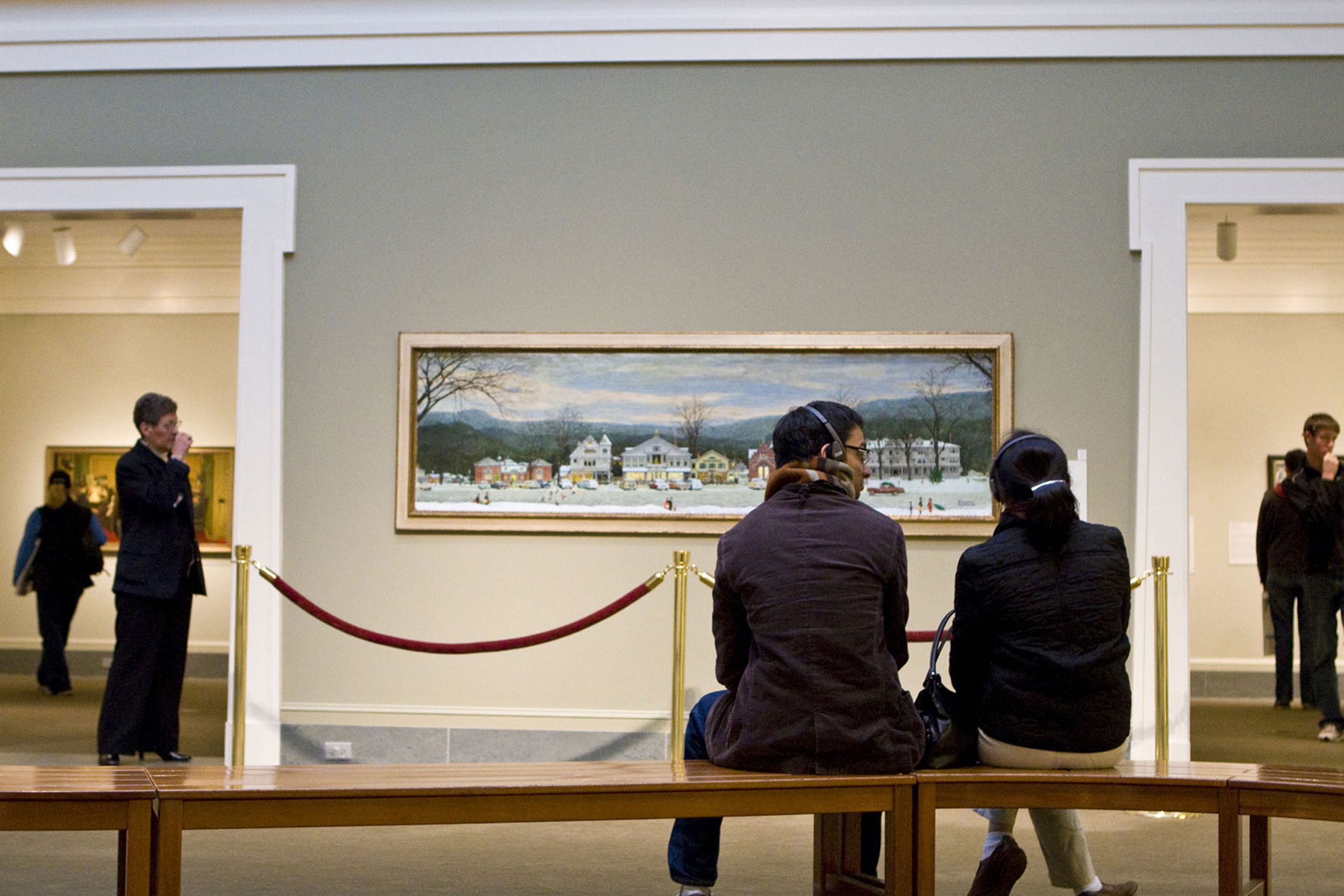 Two guests wearing audio headphones sit on a bench in front of Norman Rockwell's, 'Home for Christmas (Stockbridge Main Street at Christmas).'
