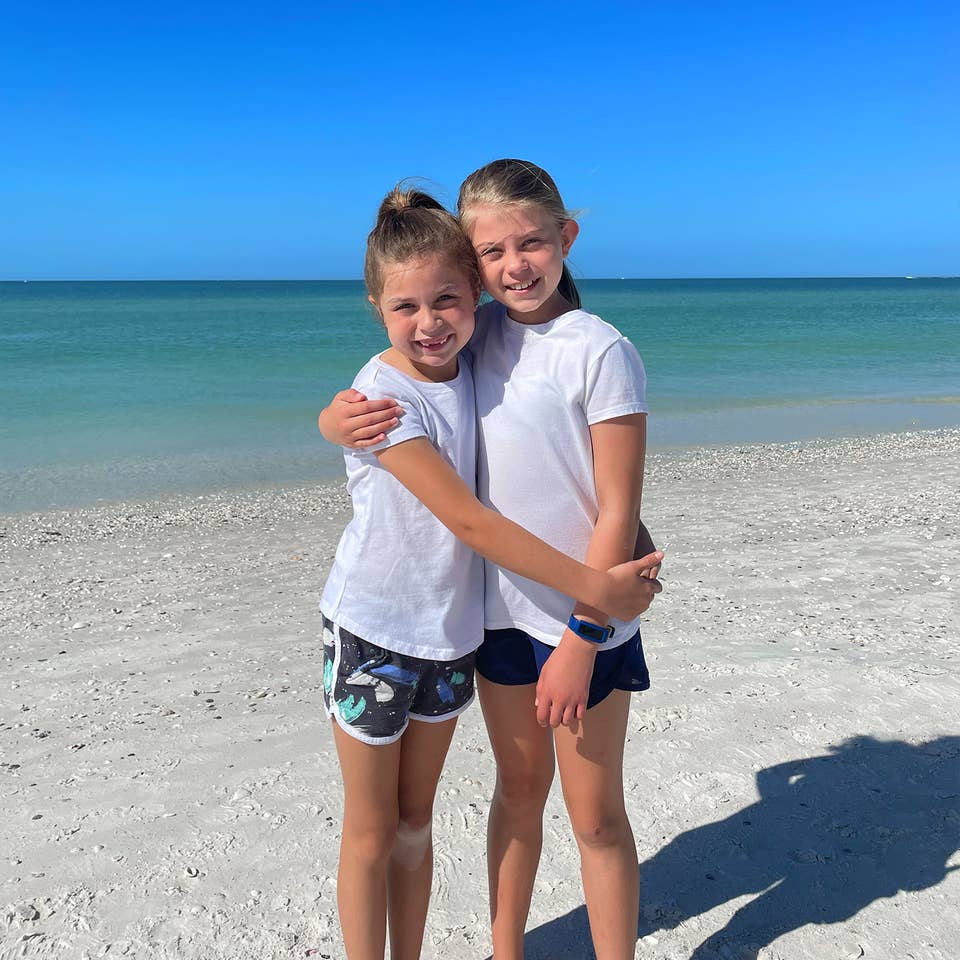 Two caucasian girls wearing white t-shirts and running shorts stand on a beach.