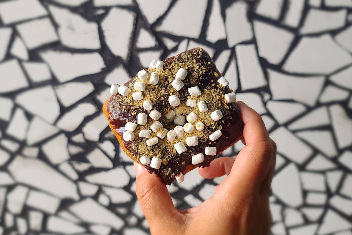 A hand holds a square-shaped doughnut near a white mosaic tile coated with mini marshmallows, chocolate and graham cracker dust.