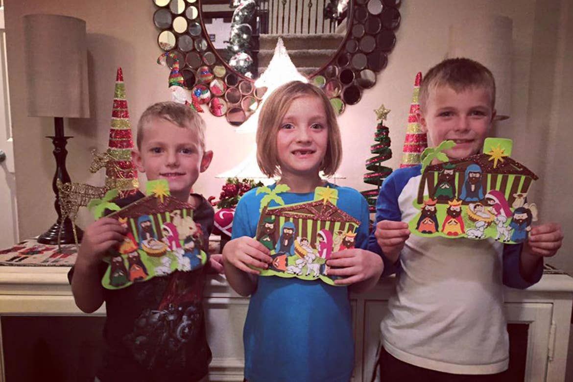 Three children hold up their crafted paper Nativity scenes in front of some holiday decorations.