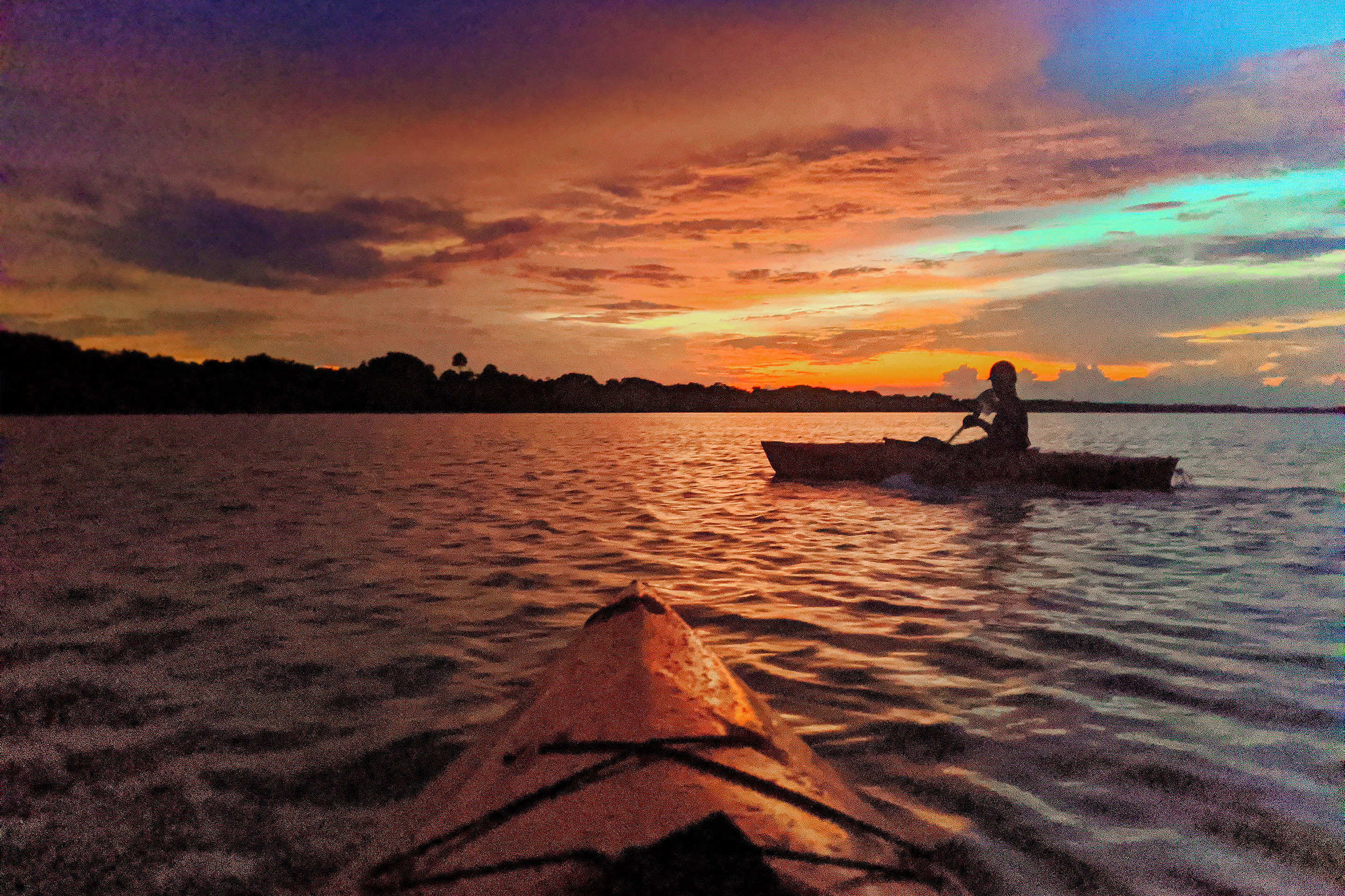 A man on a red kayak paddles under a sunset in the Mosquito Lagoon at Merritt Island National Wildlife Refuge where bioluminescence glows.