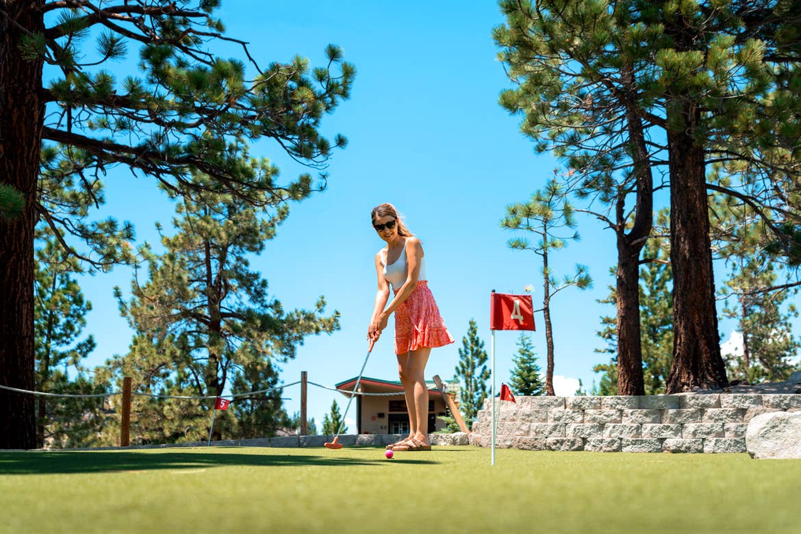 A caucasian woman wearing a white tank top and red, patterned skirt with sunglasses holds a putter on a green outdoors under a blue sky surrounded by various pine trees.
