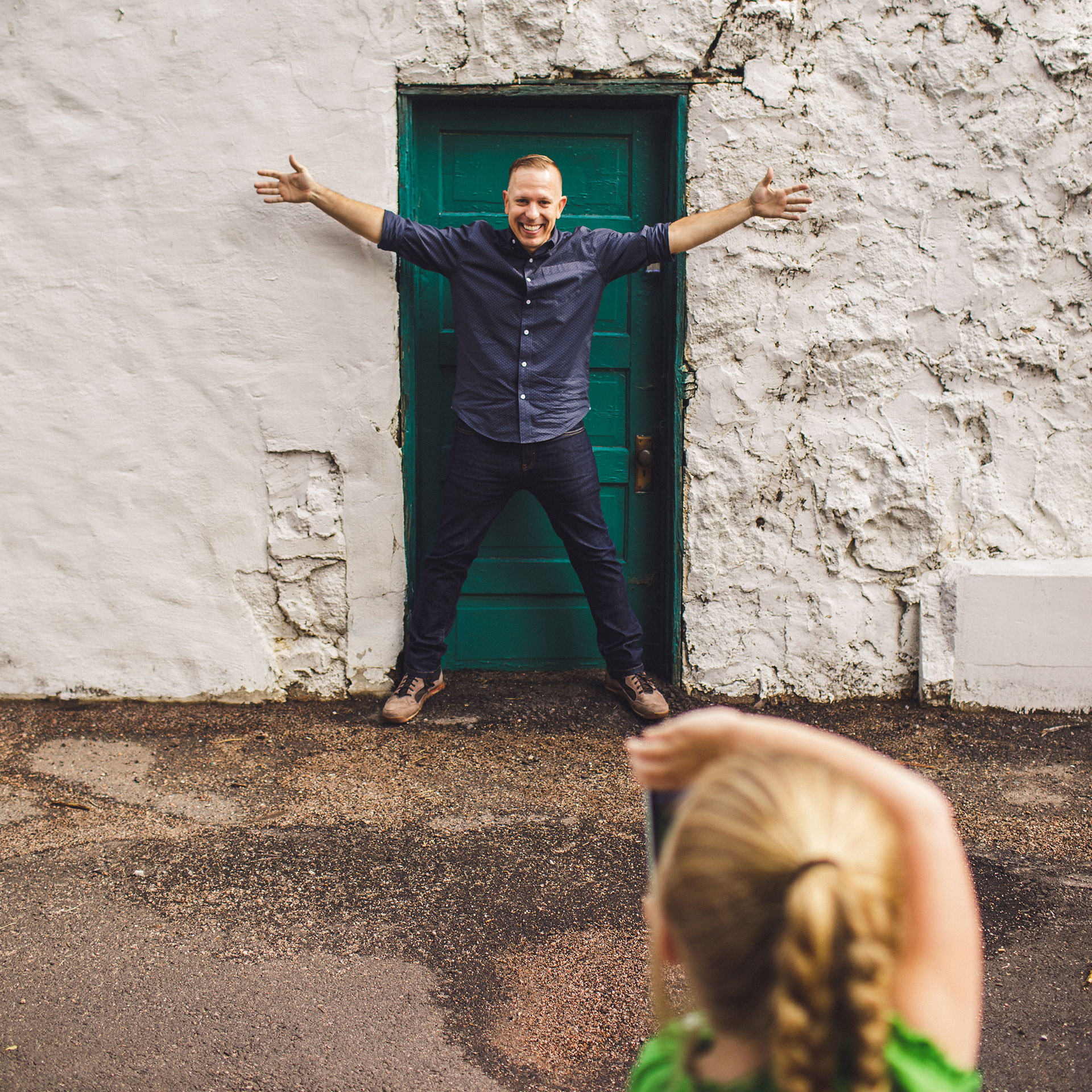 A father stands in front of a green door posing excitedly while his daughter takes his photograph.