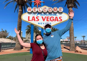 Featured Contributor, Ashley Fraboni (left) and her fiancé, Nicholas (right), pose in front of the 'Welcome to Las Vegas' marquee while wearing face masks.
