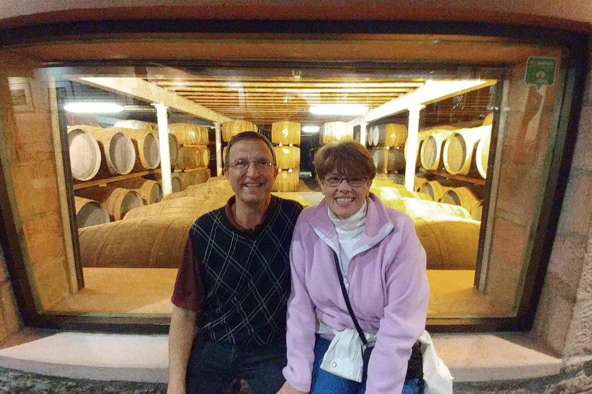 A caucasian male wearing a dark patterned polo (left) and a caucasian woman wearing a pink fleece jacket and bag (right) sit outside a window of a distillery in Inverness, Scotland.