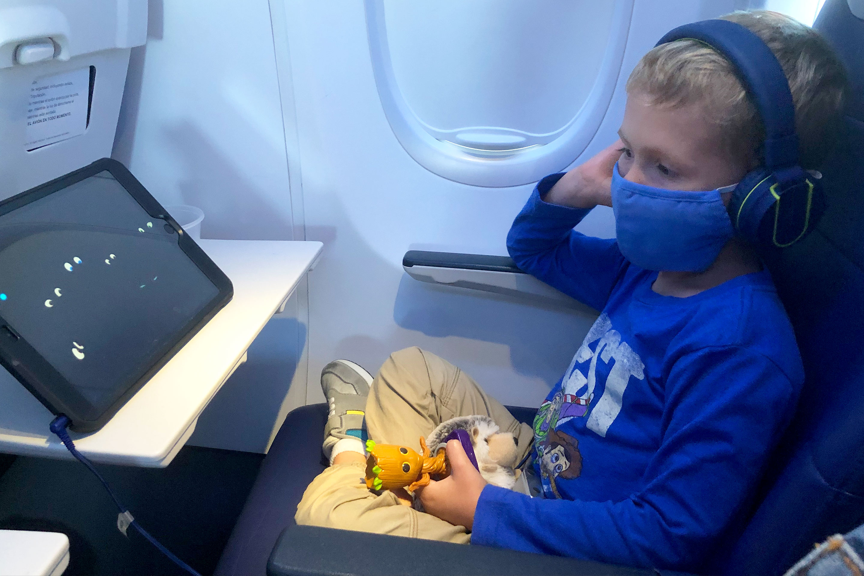 Sarah's son, Logan, sits on the airplane with his mask and entertainment on the tray.