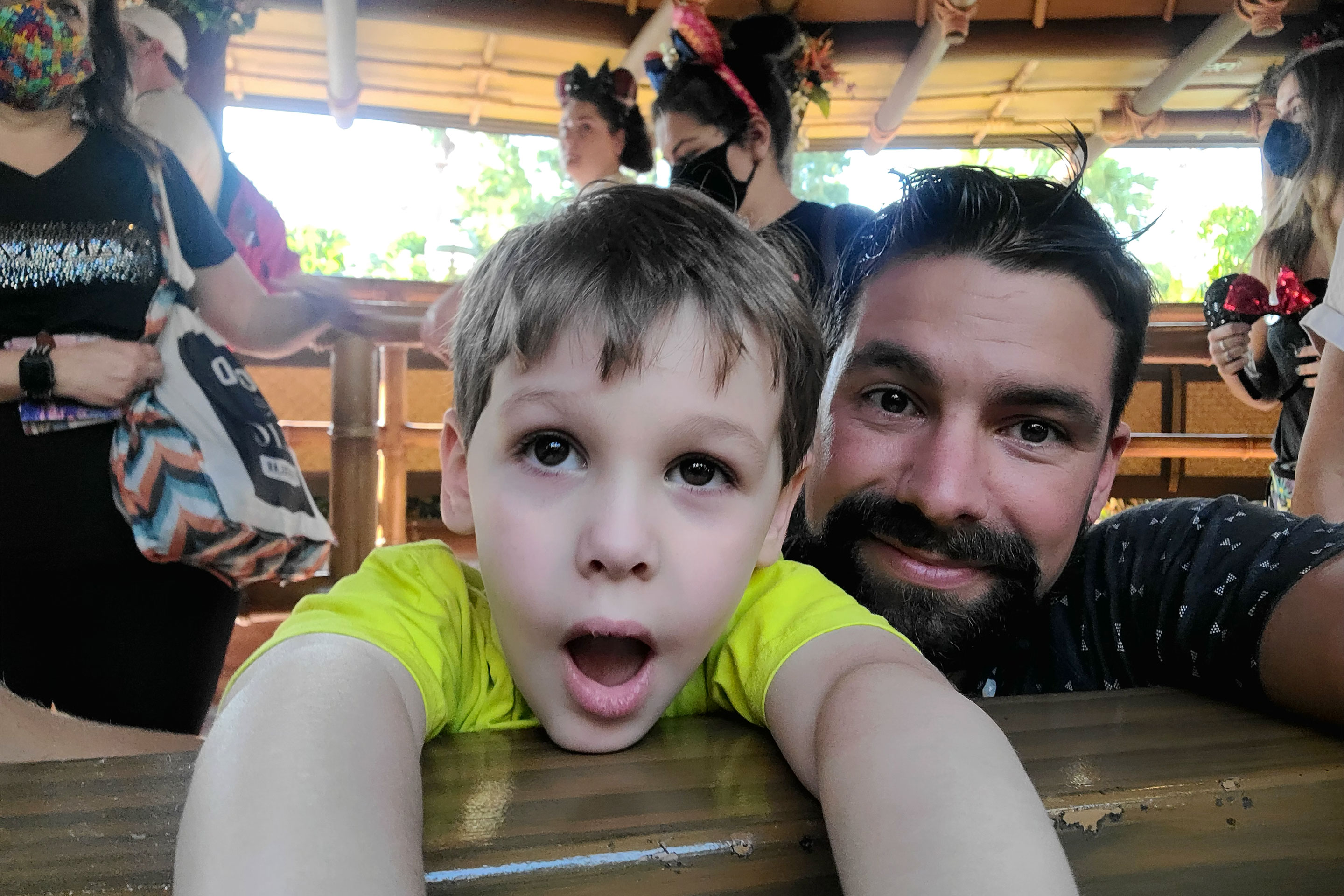A young boy (left) and man (right) stand in an attraction queue surrounded by guests in safety masks and mouse ears.
